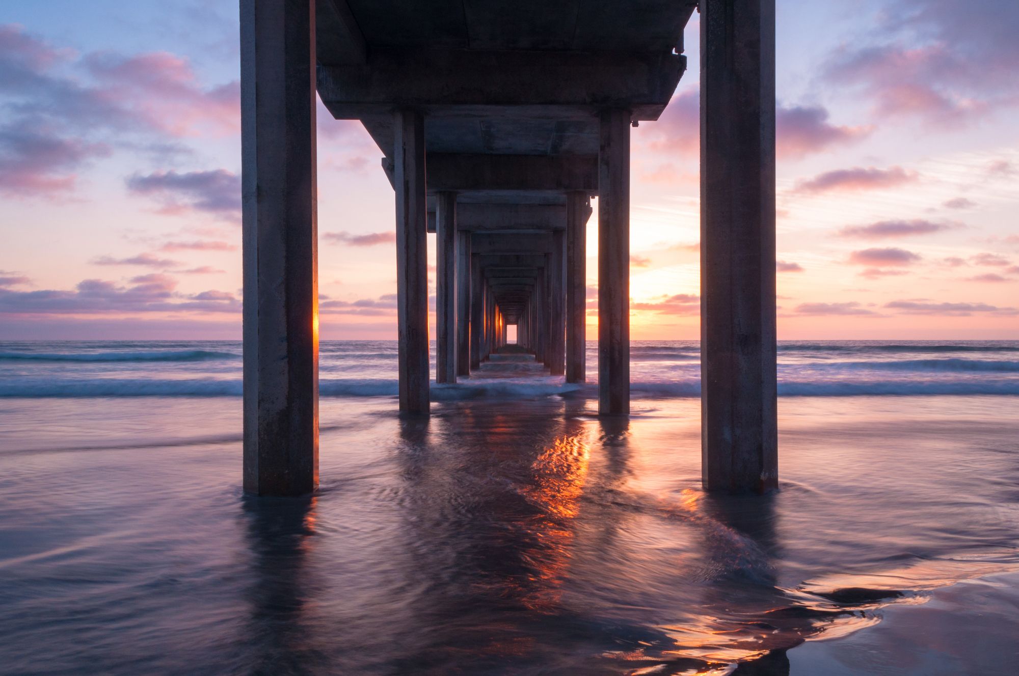 Long-exposure sunset under a La Jolla pier, the sun's reflection streaking down the passageway between concrete columns